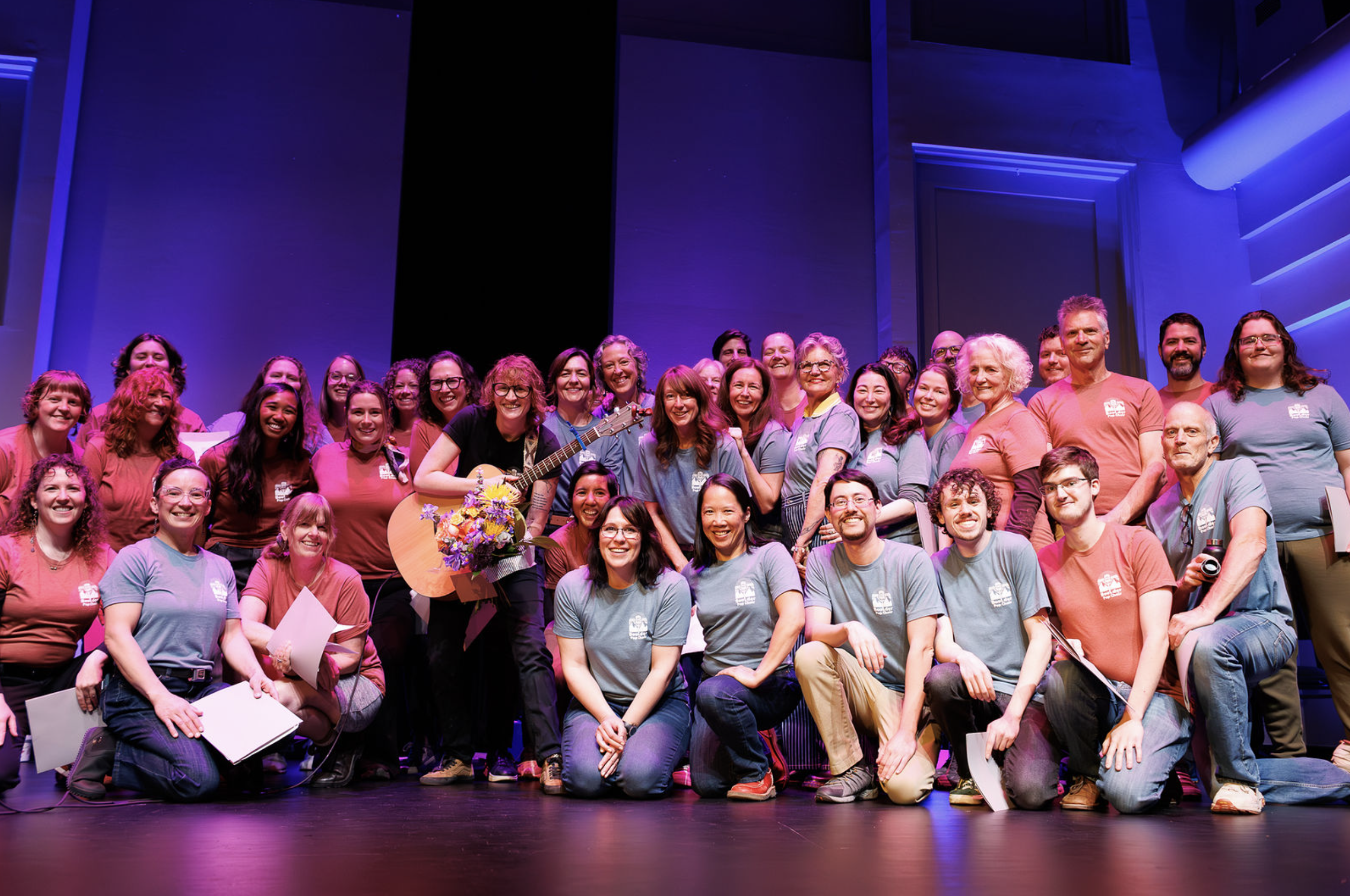 The full Boulder Pop Choir smiling together on stage in matching red and blue shirts, with their guitarist holding a bouquet of flowers.