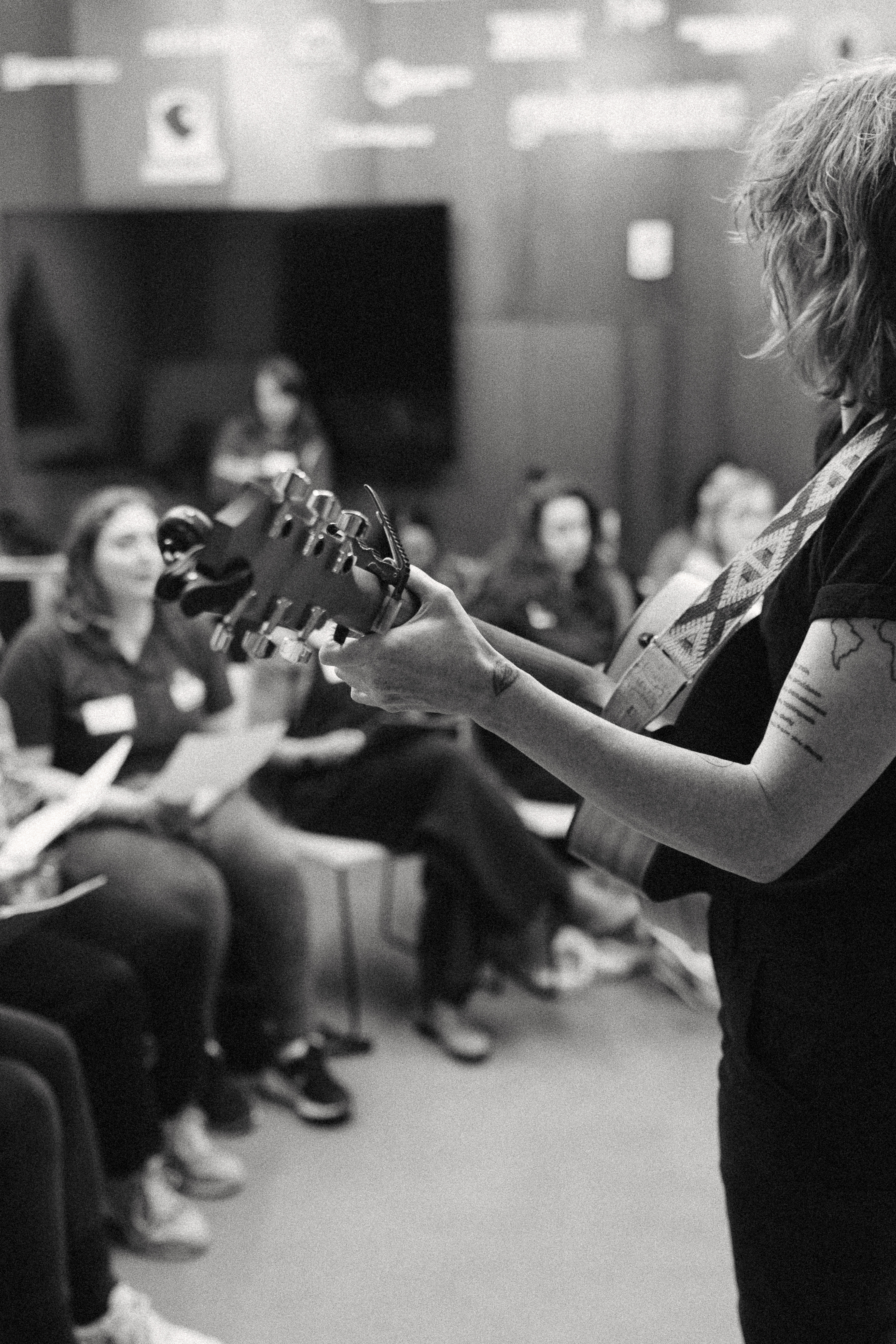 Close-up of hands playing guitar at rehearsal, with choir members seated and blurred in the background, in black and white.
