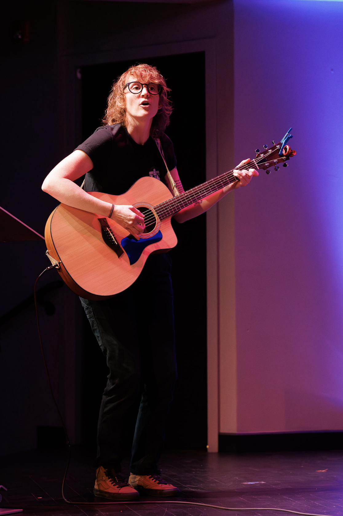 The choir's guitarist singing and playing an acoustic guitar on stage under purple light.