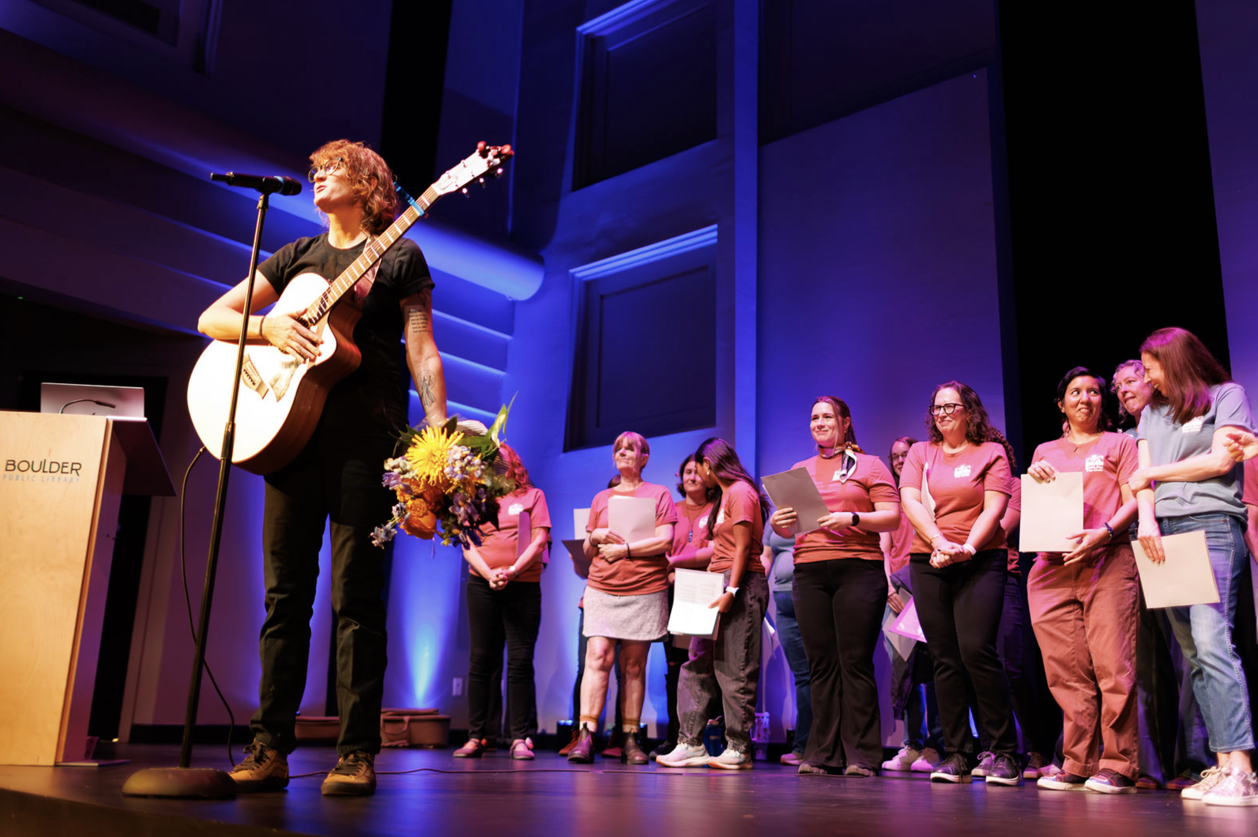 The choir standing on stage under blue and purple lights, holding sheet music, while the guitarist speaks at a microphone.